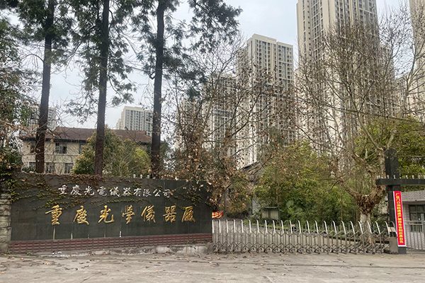 The factory gate at Xiema: a dark stone wall with gold characters naming Chongqing Optical and Electrical Instrument Co., Ltd and Chongqing Optical Instrument Factory, trees behind